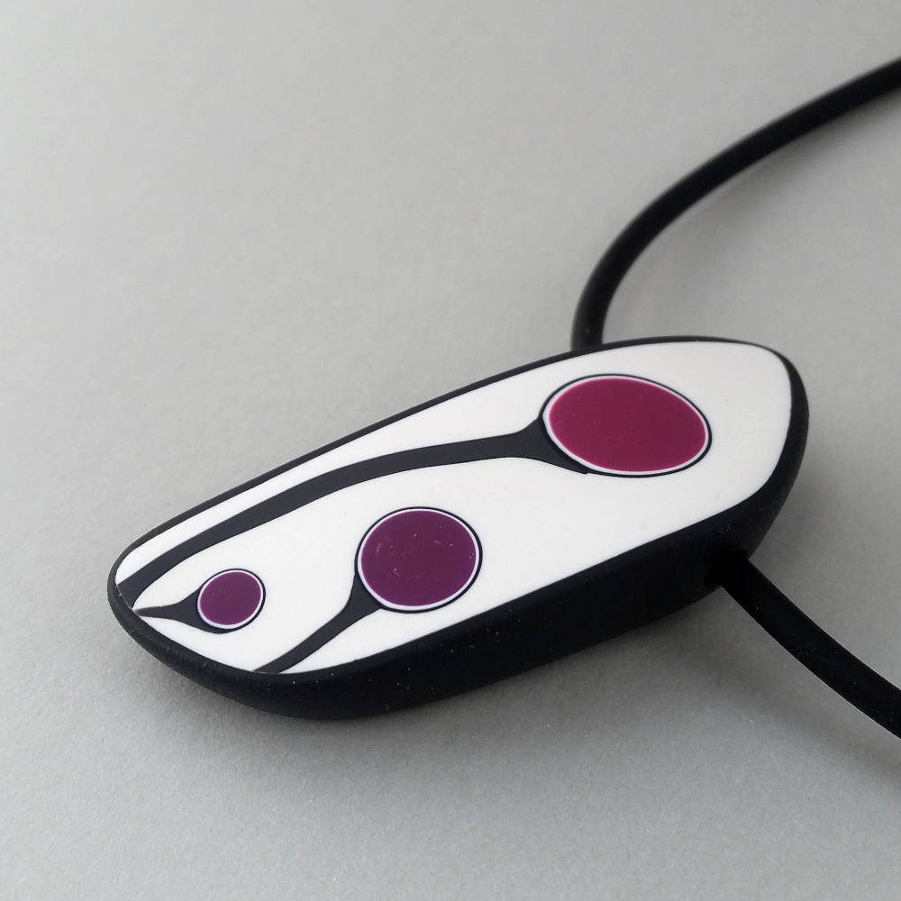 Handmade shield shaped necklace featuring an abstract flower bud motif in plum on a white background, with charcoal border. It is approximately 2.6 cm wide and 6. long and hangs on a black adjustable cord.